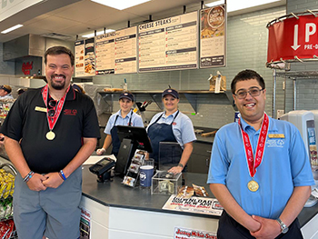 Special Olympics athletes wearing gold medals stand on either end of the counter at a Jersey Mike's location while two Jersey Mike's employees stand behind them in front of the menu board.