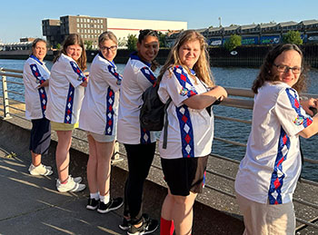 Six women in Special Olympics USA team shirts stand along a rail over a river and look back at the camera over their right shoulders.