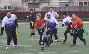 Young football player in an orange jersey runs right to left, trying to outrun several defenders.