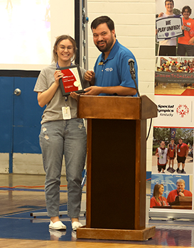 A woman in a gray t=shirt and jeans stands to the left accepting a plaque from a man in a blue shirt who stands behind a podium.