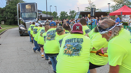 A line of men in bright yellow shirts pull a UPS 18-wheeler.