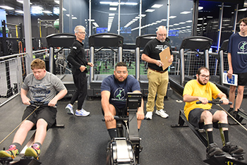 Athletes on rowing machines while scorekeepers watch behind them.