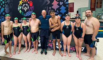 Swimmers in suits and caps surround Olympics gold medalist and swimming commentator Rowdy Gaines. 