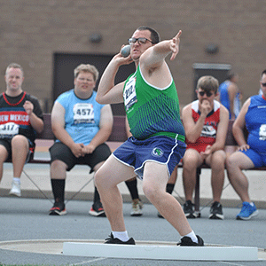 Man in a green and blue track uniform competes in the shot put.